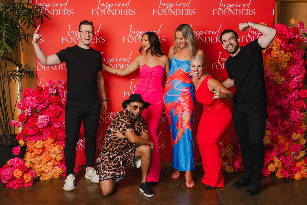 Six guests striking playful poses against the red Inspired Founders backdrop